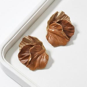 Può includere: Due orecchini a forma di fiore con un design a petali stratificati. Gli orecchini presentano una sfumatura di colori, che passa da una tonalità dorata nella parte superiore a un marrone intenso alla base. Sono posizionati su uno sfondo bianco all'interno di un vassoio bianco.