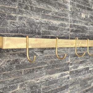 Könnte beinhalten: Ein goldfarbenes Metallregal mit sechs Haken, das an einer grauen Steinwand montiert ist. Das Regal ist für Küchenutensilien oder andere Gegenstände gedacht.