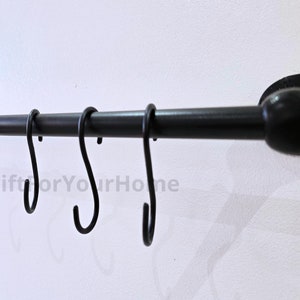 Puede incluir: Un estante de utensilios de cocina de metal negro con seis ganchos montado en una pared blanca. El estante tiene un remate redondo en el lado derecho.