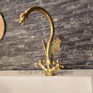 May include: A gold swan-shaped faucet with two handles. The faucet is mounted on a white countertop.