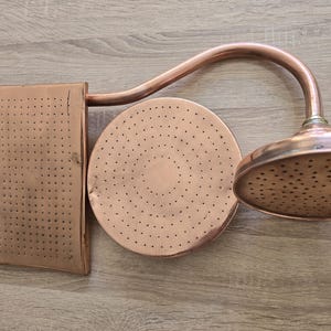 May include: A copper shower head set, featuring a rectangular wall mount, a circular shower head, and a curved pipe. The set has a vintage aesthetic with numerous small holes for water distribution. The copper has a warm, reddish-brown hue.