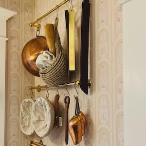 Peut inclure: Un support de rangement de cuisine avec divers articles suspendus. On y trouve une casserole en cuivre, un panier tressé, des ustensiles en bois et une sangle marron foncé. Le support est en métal doré et est fixé sur un mur à motifs.