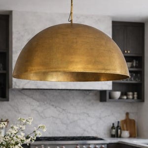 May include: A kitchen interior featuring a large, gold-colored dome pendant light. The kitchen island has a dark countertop with a wooden cutting board, a bowl of lemons, and a vase of white flowers. Dark wood cabinets and a stainless steel stove are visible.