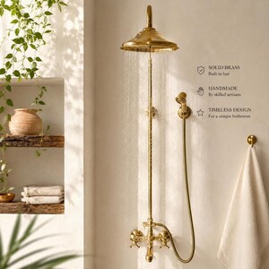 Könnte beinhalten: Ein goldfarbenes Duschsystem aus Messing mit einem großen runden Regenduschkopf, einem Handbrauseaufsatz und Kreuzgriffventilen. Das System sprüht Wasser. Der Text an der Wand lautet: SOLID BRASS, HANDMADE, TIMELESS DESIGN.