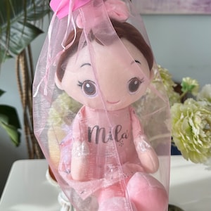 Puede incluir: Una mu&ntilde;eca de peluche rosa con cabello casta&ntilde;o y grandes ojos marrones, que lleva un vestido rosa con el nombre "Mila" bordado. La mu&ntilde;eca est&aacute; envuelta en una bolsa de tul rosa con un lazo rosa.