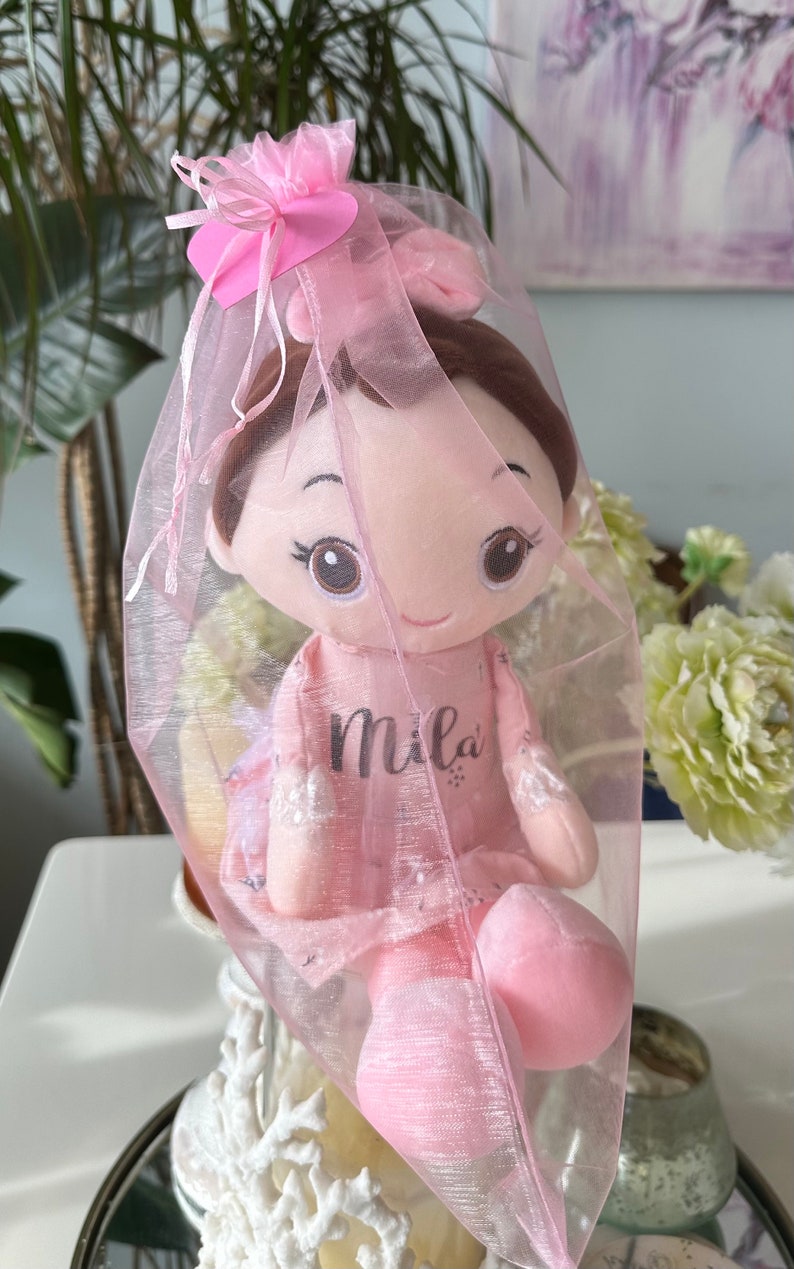 Puede incluir: Una mu&ntilde;eca de peluche rosa con cabello casta&ntilde;o y grandes ojos marrones, que lleva un vestido rosa con el nombre "Mila" bordado. La mu&ntilde;eca est&aacute; envuelta en una bolsa de tul rosa con un lazo rosa.