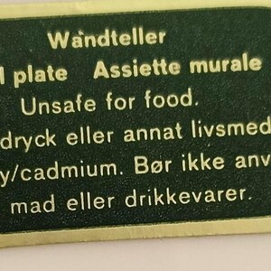 May include: A dark green, rounded rectangular label with a gold border and gold text. The text reads: Wandteller, Wall plate, Assiette murale, Unsafe for food, Ej f&ouml;r dryck eller annat livsmedel, iver bly/cadmium, B&oslash;r ikke anvendes til mad eller drikkevarer.