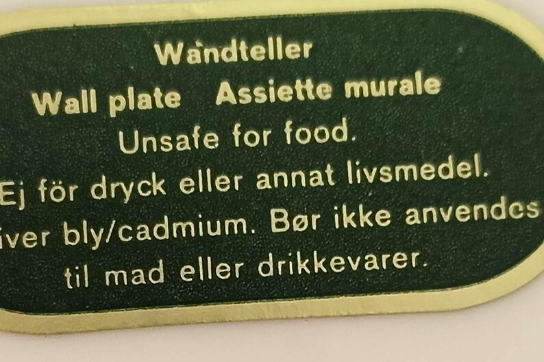 May include: A dark green, rounded rectangular label with a gold border and gold text. The text reads: Wandteller, Wall plate, Assiette murale, Unsafe for food, Ej f&ouml;r dryck eller annat livsmedel, iver bly/cadmium, B&oslash;r ikke anvendes til mad eller drikkevarer.
