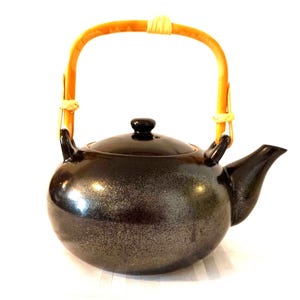 Op de afbeelding: Een zwarte theepot van keramiek met een gespikkelde afwerking en een bamboe handvat. De theepot heeft een deksel en een tuit.