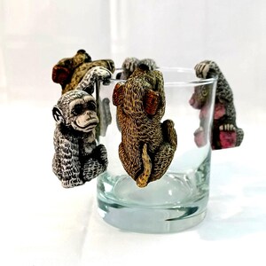 Puede incluir: Cuatro pequeñas figuritas de animales, un mono, un perro y dos osos, cuelgan del borde de un vaso transparente. El mono es gris y blanco, el perro es marrón y blanco, y los osos son marrones y rosas.