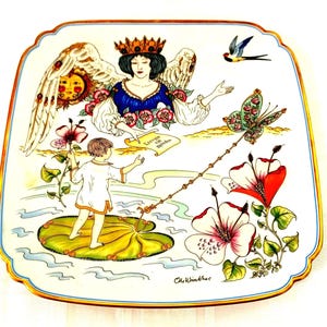 May include: A square decorative porcelain plate with a gold-trimmed edge featuring a colorful illustration of a winged figure wearing a crown, a child standing on a lily pad, large hibiscus flowers, a butterfly, and a swallow with the text Eervia kai Agapes.