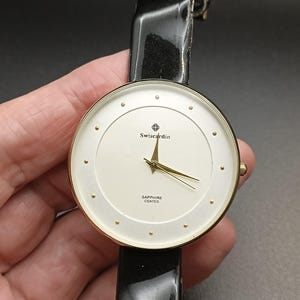 May include: A gold-tone wristwatch with a white face and black leather band. The watch face features the brand name "Swisscardin" and the words "Sapphire Coated".