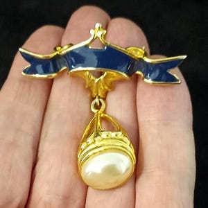 Puede incluir: Broche dorada con un diseño de cinta esmaltada azul marino y un colgante en forma de lágrima. El colgante tiene un marco dorado y una gran perla ovalada de color crema. Accesorio vintage.