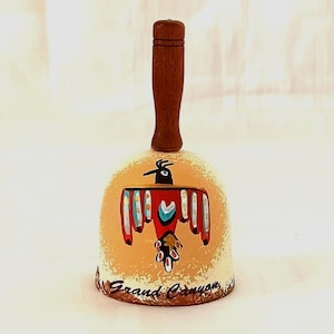 Peut inclure: Une petite cloche en céramique avec une poignée en bois. La cloche est peinte avec un motif d'oiseau stylisé en rouge, jaune, bleu et noir. Les mots "Grand Canyon" sont écrits au bas de la cloche.