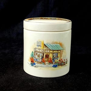 May include: White ceramic jar with a lid featuring a painted image of a small shop with a green roof and red flowers. The shop has a sign that reads "Post Office".