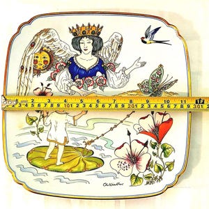 May include: A square ceramic plate with rounded corners featuring a colorful illustration of a crowned angel, a child on a lily pad, a sun, a butterfly, and flowers. A yellow measuring tape shows the plate is approximately 12 inches wide.
