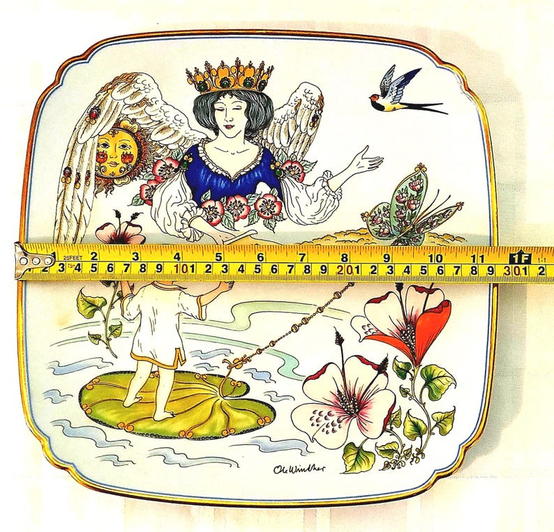 May include: A square ceramic plate with rounded corners featuring a colorful illustration of a crowned angel, a child on a lily pad, a sun, a butterfly, and flowers. A yellow measuring tape shows the plate is approximately 12 inches wide.