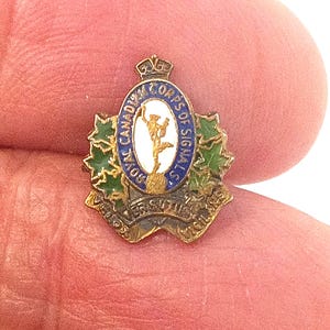 Könnte beinhalten: Ein Vintage-Abzeichen des Royal Canadian Corps of Signals. Das Abzeichen zeigt ein zentrales Emblem mit einer Figur, umgeben von einem blauen Ring mit Text. Grüne und goldene Akzente sind vorhanden, mit zusätzlichem Text am unteren Rand. Das Abzeichen wird in einer Hand gehalten.
