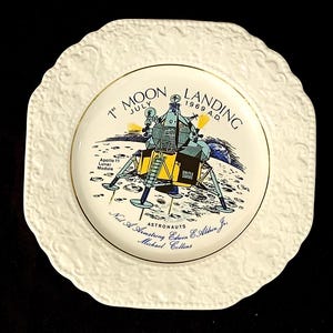 May include: A decorative plate commemorating the 1969 Apollo 11 moon landing. The plate features a detailed illustration of the lunar module on the moon's surface, with text including the date and astronaut names. The plate has a gold rim and a white, embossed border.