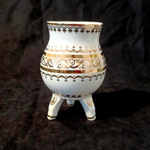 May include: A white ceramic vessel with a rounded body and three short legs. The exterior features gold-coloured decorative bands with intricate scrollwork and geometric patterns around the rim and the centre of the piece, creating a contrast.