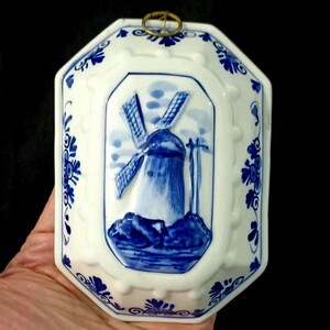 May include: White ceramic wall hanging with a blue windmill design. The octagonal piece features a raised windmill scene and blue floral accents. A brass ring is attached at the top for hanging. The piece is held by a hand.