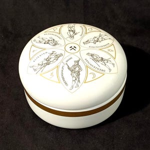 May include: White ceramic round box with a gold trim and a design of a pickaxe and six figures in traditional German clothing. The figures are labeled with German words, including "Bergmuskat", "Altester", "Oberberghauptmann", "Oberzehnter", "Marktscheuer", and "Bester Mann".