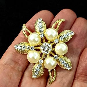 Puede incluir: Broche floral dorado con cinco grandes perlas de imitación y numerosos cristales transparentes. El broche tiene un diseño detallado y texturizado con un grupo central de cristales y detalles de perlas.