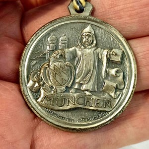 Puede incluir: Un medallón de metal plateado con una imagen en relieve de una persona con una túnica que sostiene un libro y un escudo con un león. El medallón está inscrito con la palabra "MUNCHEN" y el texto "DESCHLER MUNCHEN 90".