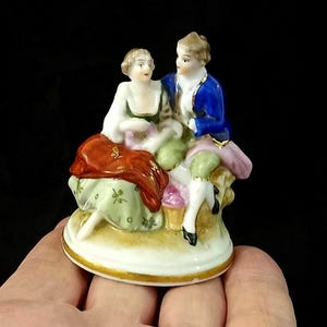May include: A small porcelain figurine depicting a couple in period clothing. The man wears a blue jacket and the woman a white dress with a red shawl. The base is white with gold trim, and the figures are in a seated pose.