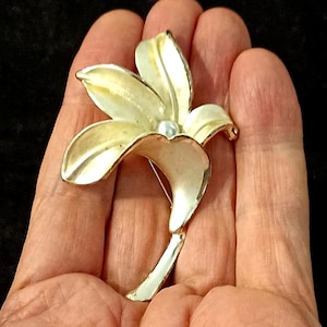 Puede incluir: Broche de flores plateado con centro de perla. El broche presenta cinco pétalos con un acabado amarillo claro y blanco. El broche se sostiene en una mano sobre un fondo negro.