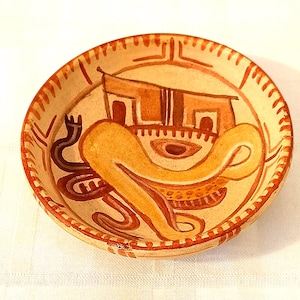 May include: A small, round, hand-painted ceramic bowl with a cream-colored base. The bowl features a stylized design in shades of brown and orange, including a house-like structure and abstract shapes. The rim is decorated with a repeating pattern.