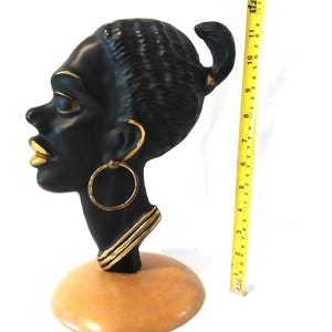 Puede incluir: Escultura de cerámica negra de la cabeza de una mujer con detalles dorados. La escultura está sobre una base de madera.