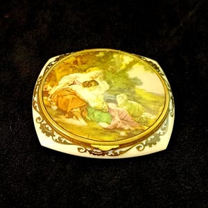 Op de afbeelding: Een wit porseleinen compact met een gouden rand en een geschilderd tafereel van mensen in een landschap. De compact heeft een scharnierend deksel.
