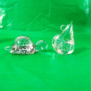 Puede incluir: Dos figuritas de vidrio transparente, una con forma de tortuga y la otra con forma de lágrima, ambas con burbujas de aire atrapadas en su interior.
