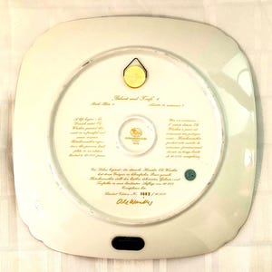 May include: A square white porcelain decorative plate with rounded corners, featuring gold text on the back. The text includes 'Geburt und Taufe 1', 'Birth Plate 1', and 'Limited Edition No. 3882 / 10.000'. A gold metal hanger is attached to the top.