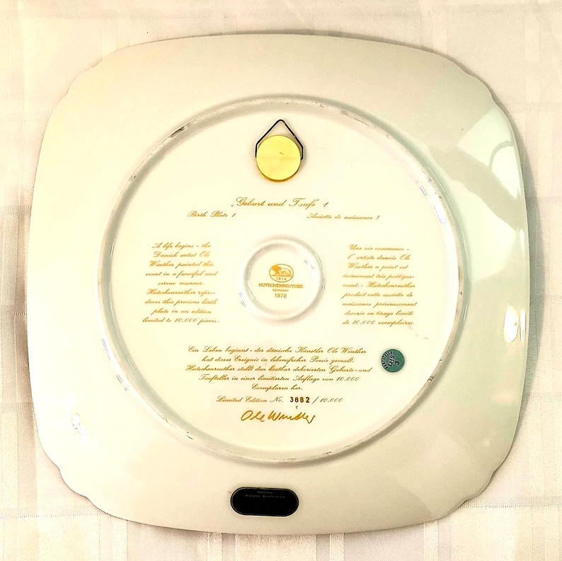 May include: A square white porcelain decorative plate with rounded corners, featuring gold text on the back. The text includes 'Geburt und Taufe 1', 'Birth Plate 1', and 'Limited Edition No. 3882 / 10.000'. A gold metal hanger is attached to the top.