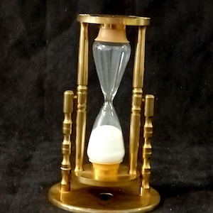 May include: An hourglass featuring a polished brass frame with vertical supports and a circular base. The clear glass bulb contains white sand, with the majority of the sand collected in the bottom chamber, held by light tan plastic caps.