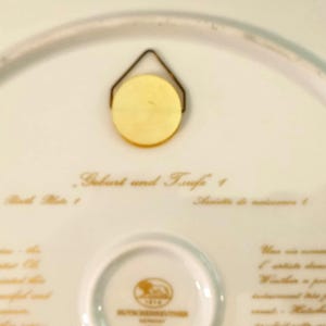 May include: The underside of a white porcelain plate featuring a gold-colored metal wall hanger. The text reads Geburt und Taufe 1, Birth Plate 1, and Assiette de naissance 1, with a central Hutschenreuther logo and additional printed text.