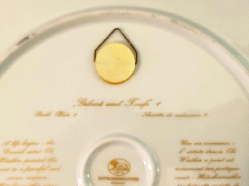 May include: The underside of a white porcelain plate featuring a gold-colored metal wall hanger. The text reads Geburt und Taufe 1, Birth Plate 1, and Assiette de naissance 1, with a central Hutschenreuther logo and additional printed text.