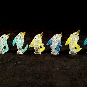 May include: A set of six glass dolphin figurines in various colours, including blue, yellow, and white. The dolphins are all facing the same direction and have a textured, bubbly surface.