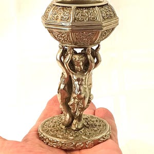 May include: A silver-toned metal candle holder featuring a bowl with intricate embossed floral patterns supported by a central pillar of three sculpted figures standing on a circular base with matching embossed decorative detailing.