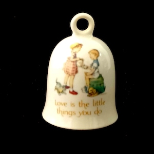 May include: White porcelain bell with a handle. The bell features a colourful illustration of two children and a dog. The text "Love is the little things you do" is printed on the bell.