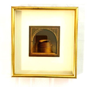 May include: A square framed artwork featuring a gold-toned metallic frame and a white mount. The central image depicts a dark brown, arched architectural niche containing a cylindrical, tiered object with a textured surface in shades of brown.