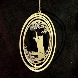 May include: Gold-coloured metal ornament featuring a silhouette of a cat reaching up to catch a dangling ball. The ornament is in the shape of an oval with a swirling design.