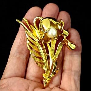 Puede incluir: Broche floral dorado con una perla en el centro. El broche tiene hojas y pétalos detallados, con un acabado texturizado. El diseño es una flor estilizada, para una chaqueta o solapa.