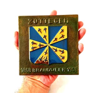 Pode incluir: Placa quadrada com a palavra "ZOTTEGEM" no topo e "1868 BRANDVEER 1968" na parte inferior. O centro apresenta um emblema azul e dourado com um design de estrela. A placa é segurada em uma mão.