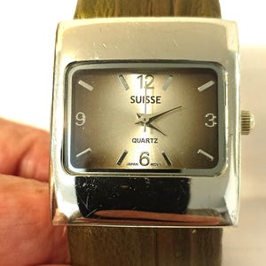 May include: A silver square-shaped wristwatch with a brown face. The watch face has black Roman numeral hour markers, black hands, and the words "SUISSE QUARTZ" and "JAPAN MOV'T". The watch has a brown leather strap.