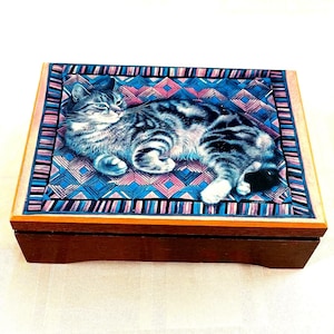 Könnte beinhalten: Eine Holzkiste mit einem Scharnierdeckel. Der Deckel zeigt ein buntes Muster mit einer grau-weißen Katze, die auf einem blauen und rosa Hintergrund liegt.