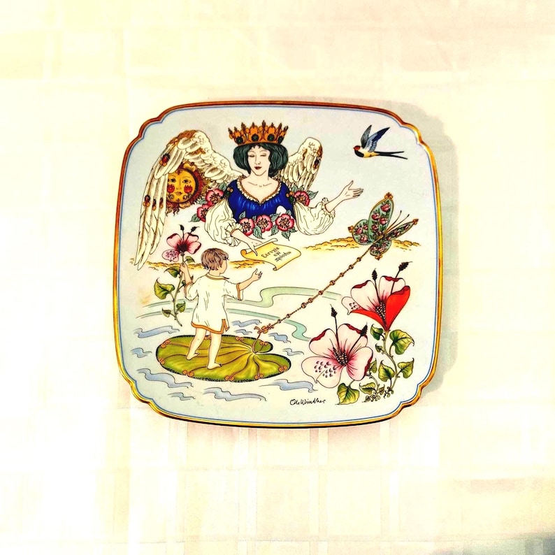 May include: A square decorative plate with rounded corners featuring a colorful illustration of a winged figure wearing a crown, a child standing on a large green leaf, a butterfly, a bird, and large flowers. The plate has a gold-toned rim.