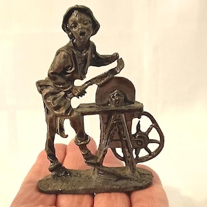 May include: A bronze figurine depicting a person sharpening a blade on a grindstone. The figure is dressed in period clothing and is working at a small, wheeled grinding machine. The figurine is held in a hand.
